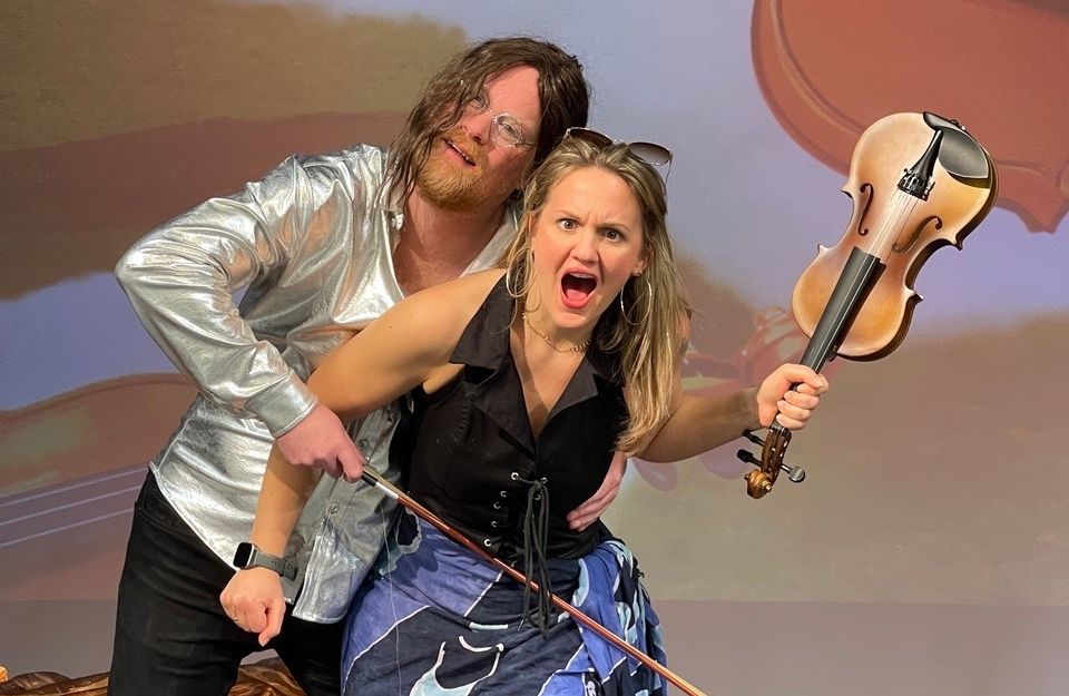 Ein Mann in silbernem Hemd und eine Frau mit Geige posieren dramatisch auf einer Bühne.