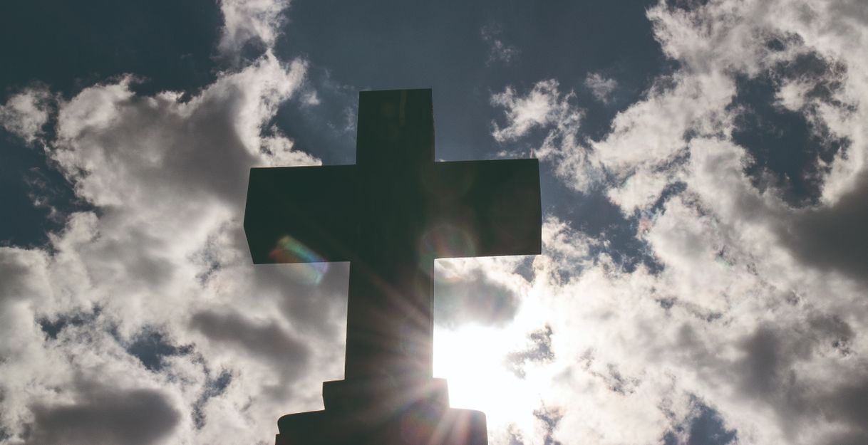 Ein Kreuz vor einem bewölkten Himmel mit Sonnenstrahlen.