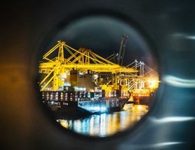 Blick durch ein Bullauge auf einen beleuchteten Hafen mit Containerschiffen und Kränen bei Nacht.