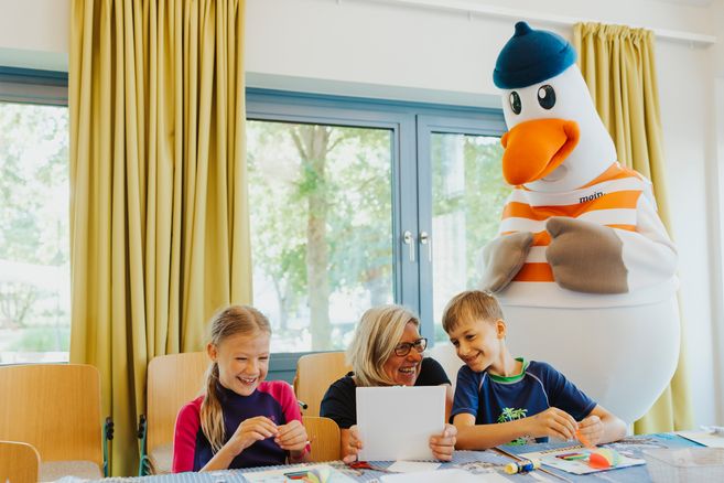 Kinder basteln mit einer Betreuerin, während ein Möwen-Maskottchen zuschaut.