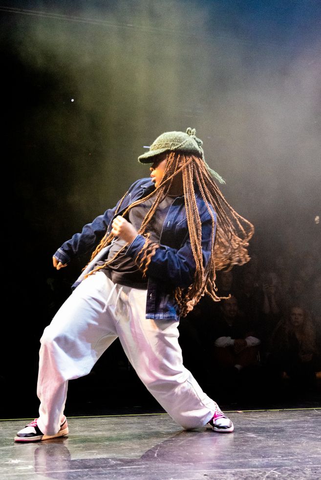 A person wearing a knitted hat, long braids, and loose clothing stands on a stage and moves their hips in one direction with bent knees. They pull themselves up in the same direction with the opposite arm.