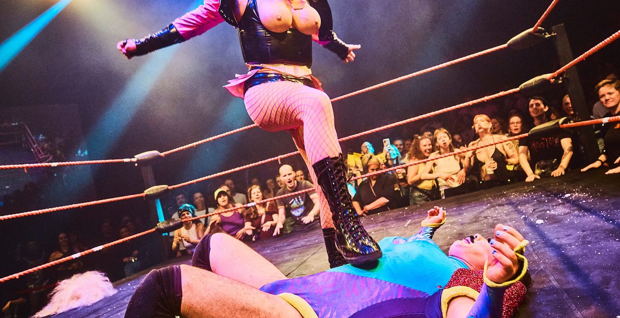 A wrestling scene: one person is lying on the floor, another person is standing over them and has their foot on the other person's chest.