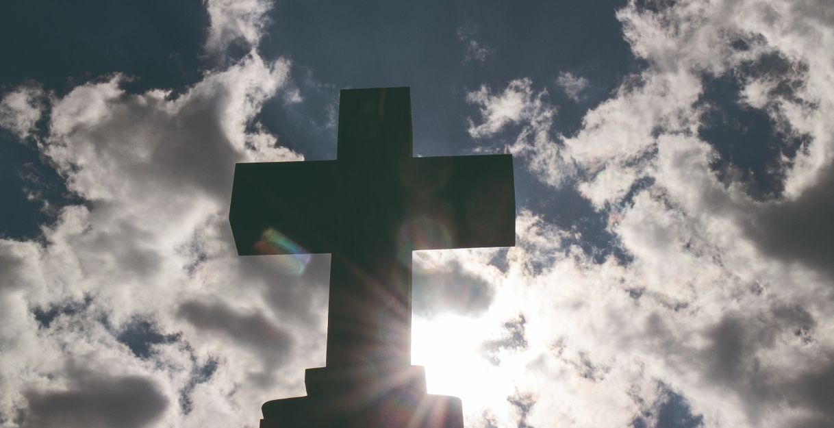 Ein Kreuz vor einem bewölkten Himmel mit Sonnenstrahlen.