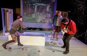 Drei Personen auf einer Bühne, eine wirft Gegenstände, eine spielt Gitarre, eine schaut zu. Hintergrund mit Projektion.