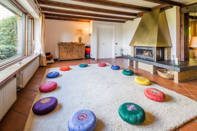 Ein gemütlicher Meditationsraum mit bunten Kissen im Kreis auf einem Teppich, einem Kamin und Holzmöbeln.