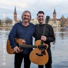 Zwei Männer mit Gitarren stehen lächelnd vor einem Fluss, im Hintergrund sind Kirchtürme zu sehen.