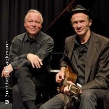 Zwei Männer sitzen vor einem Klavier, einer mit Gitarre, beide in dunkler Kleidung.