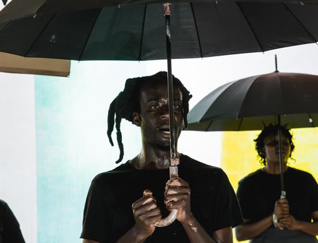 Excerpt from the production DAMBUDZO by nora chipaumire. A person stands holding an umbrella in front of a colorful wall. Behind them, two other people are visible in the background.