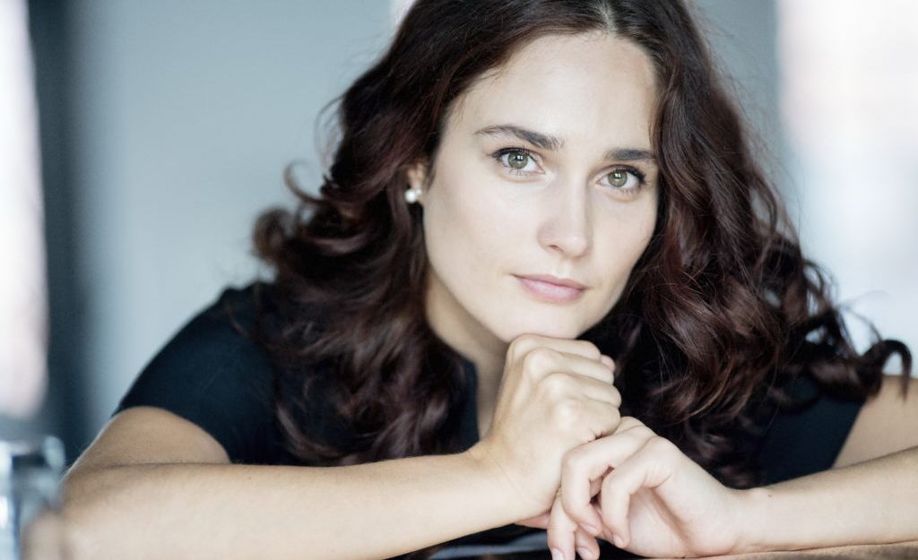 Porträt einer Frau mit langen, lockigen Haaren, die auf einem Tisch lehnt.