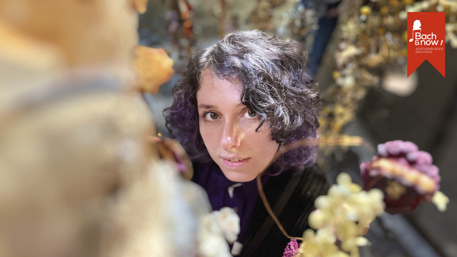 Eine Frau mit lockigem Haar schaut durch einen Blumenrahmen in die Kamera. Im Hintergrund ist ein Banner mit der Aufschrift 'Bach & now! Agathenburger Bachtage'.
