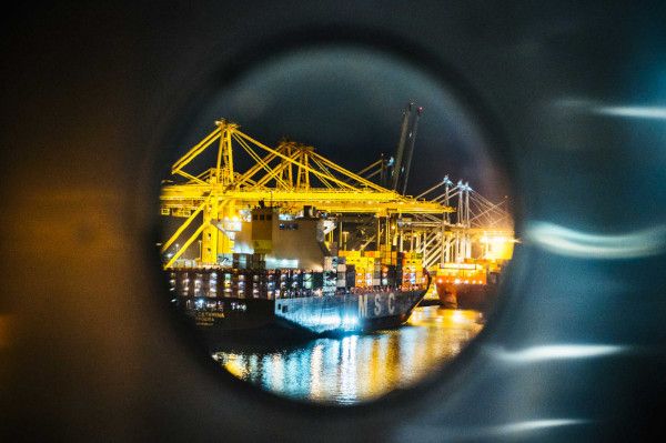 Blick durch ein Bullauge auf einen beleuchteten Hafen mit Containerschiffen und Kränen bei Nacht.