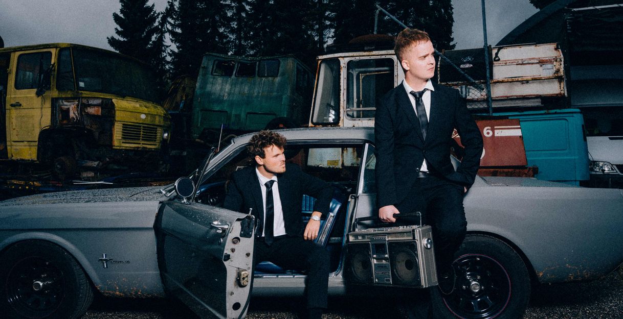 Zwei Männer in Anzügen posieren mit einem alten Auto auf einem Schrottplatz.