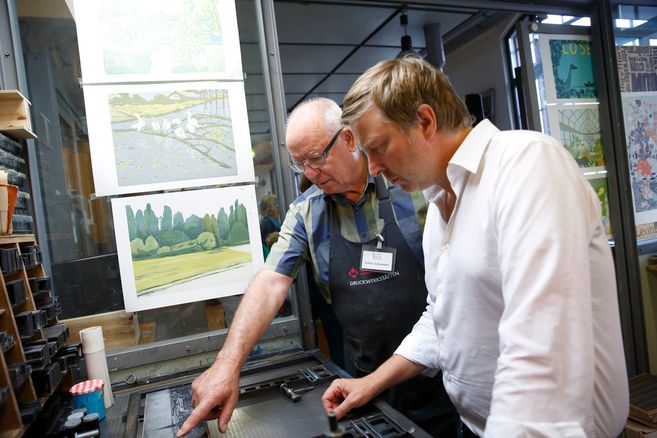 Zwei Männer arbeiten an einer Druckmaschine in einer Werkstatt mit Kunstwerken an der Wand.