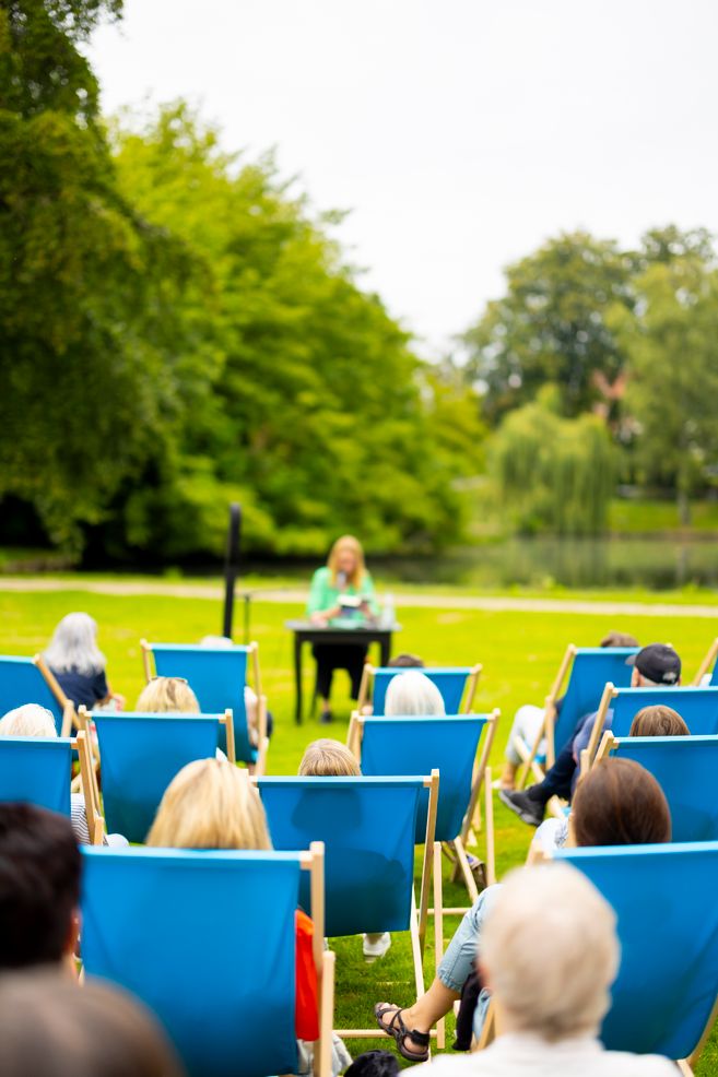 Menschen sitzen in blauen Liegestühlen in einem Park und hören einer Person an einem Tisch zu.