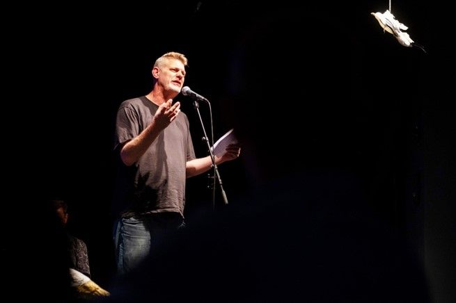 Björn Högsdal beim Poetry Slam vor einem Mikrofon, schwarzer Hintergrund, Papiervogel in der rechten Ecke