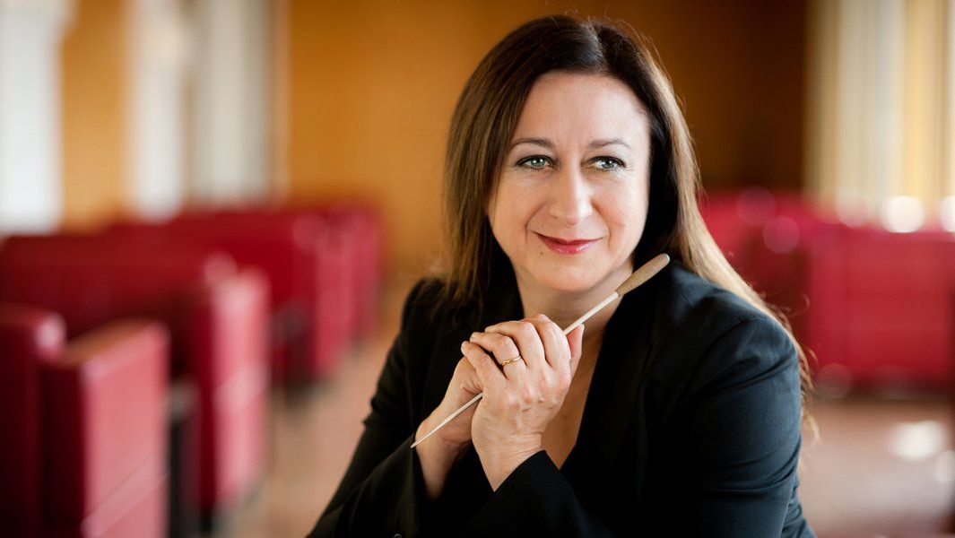 Eine Frau in schwarzer Kleidung hält einen Dirigentenstab und lächelt in die Kamera.