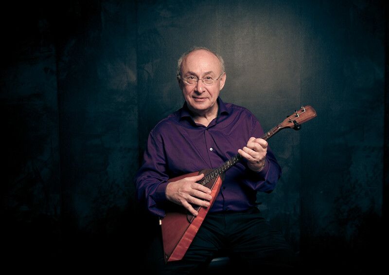 Ein Mann in einem lila Hemd spielt eine Balalaika vor einem dunklen Hintergrund.