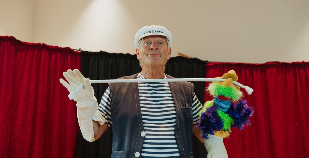 Ein Mann in Matrosenkleidung hält eine bunte Handpuppe an einem Seil vor einem roten Vorhang.