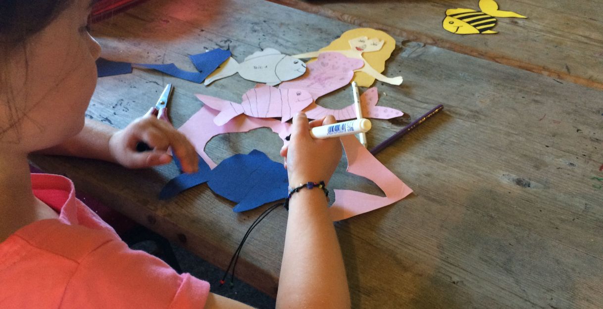 Ein Kind bastelt aus buntem Papier eine Meerjungfrau an einem Holztisch.