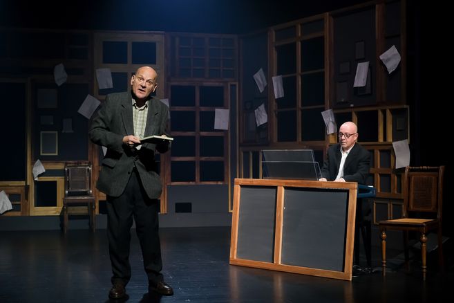 Zwei Männer auf einer Bühne, einer liest aus einem Buch, der andere spielt Klavier. Der Hintergrund ist mit Papierblättern dekoriert.