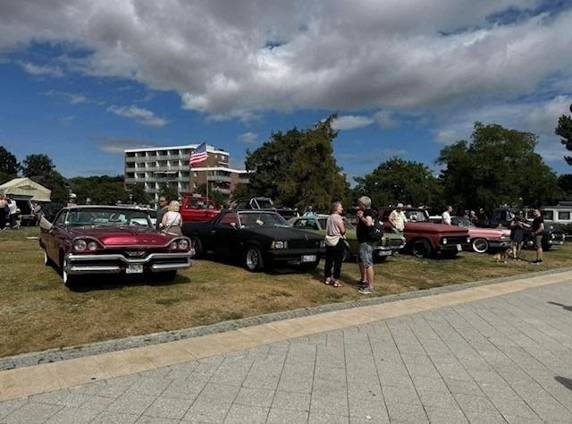 Oldtimer-Ausstellung auf einer Wiese mit mehreren klassischen Autos und Menschen, die sie betrachten. Im Hintergrund weht eine amerikanische Flagge.