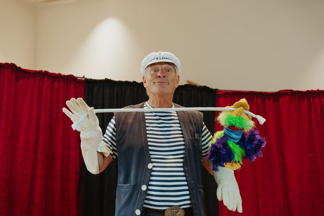 Ein Mann in Matrosenkleidung hält eine bunte Handpuppe an einem Seil vor einem roten Vorhang.