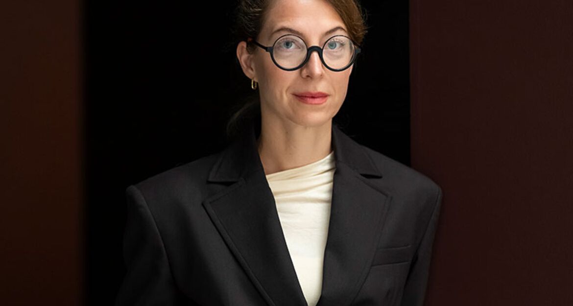 Eine Frau mit Brille und schwarzem Blazer steht vor einem dunklen Hintergrund.