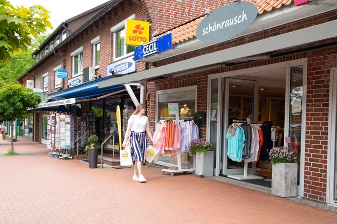 Eine Frau geht an Geschäften vorbei, darunter ein Bekleidungsgeschäft und ein Lottoladen, auf einer Einkaufsstraße.