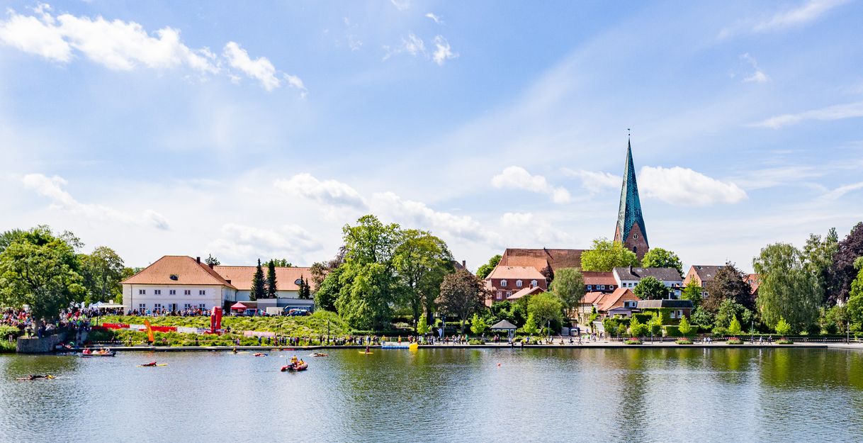 Teilnehmer des 29. Rosenstadt-Triathlons schwimmen in einem See, umgeben von malerischen Gebäuden und einer Kirche mit hohem Turm im Hintergrund.