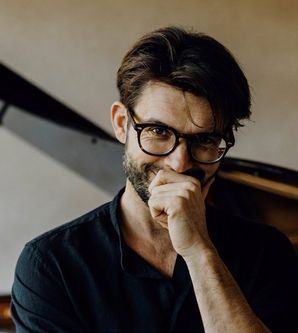 Ein Mann mit Brille steht vor einem Flügel und lächelt nachdenklich.