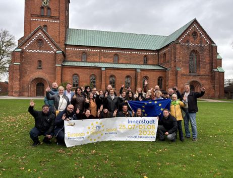 Eine Gruppe von Menschen steht vor einer Kirche und hält ein Banner hoch. Der Banner zeigt Informationen zu einer Veranstaltung in der Innenstadt.