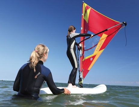 Zwei Personen beim Windsurftraining im Meer, eine steht auf dem Surfbrett mit Segel, die andere hält das Brett im Wasser.
