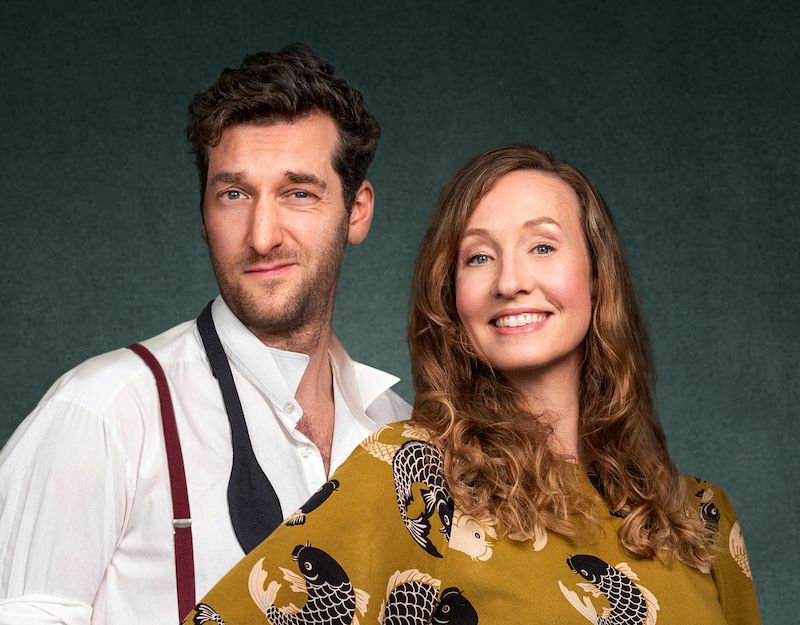 Zwei Personen posieren vor einem neutralen Hintergrund, eine Frau lächelt, ein Mann schaut ernst.