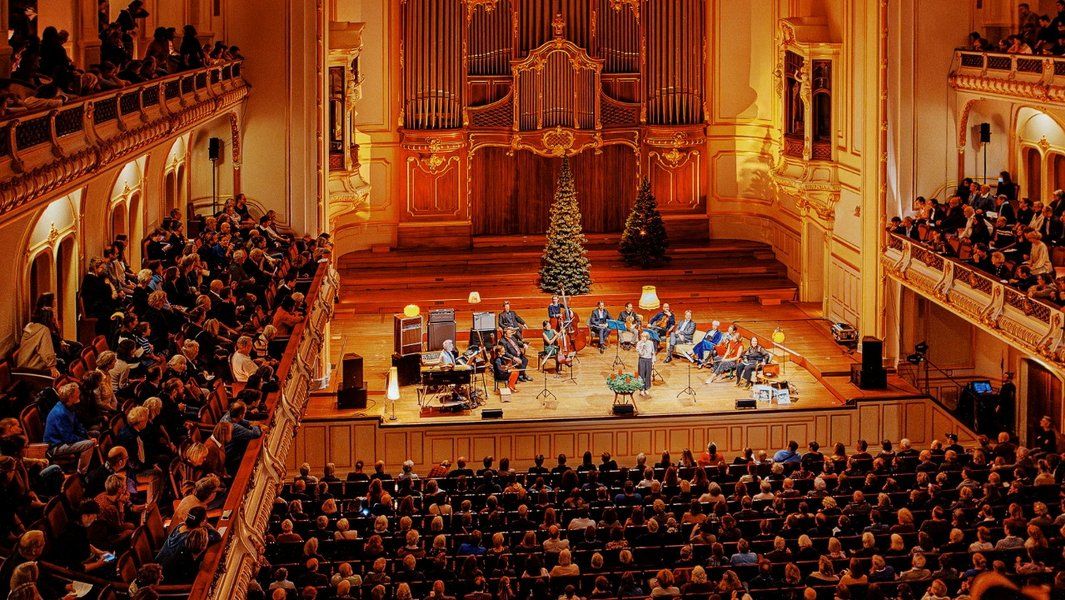 Ein Konzert in einem großen, festlich geschmückten Saal mit einem Orchester auf der Bühne und einem vollen Publikum.