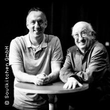 Zwei Männer stehen an einem runden Tisch und lächeln in die Kamera.