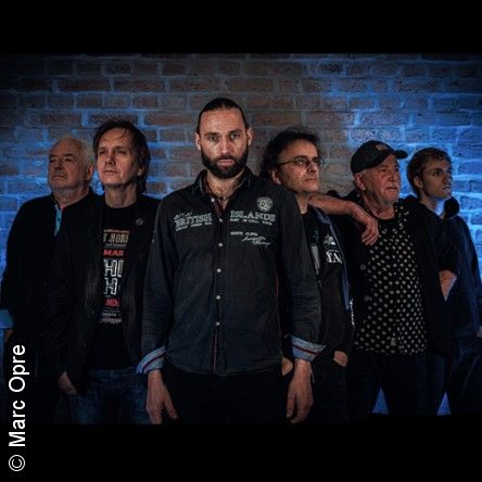 Eine Gruppe von sechs Männern steht vor einer Backsteinwand, einige schauen in die Kamera, andere zur Seite. Die Beleuchtung ist blau.