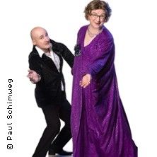 Ein Paar in eleganter Kleidung posiert fröhlich. Der Mann trägt einen schwarzen Anzug, die Frau ein lila Kleid.