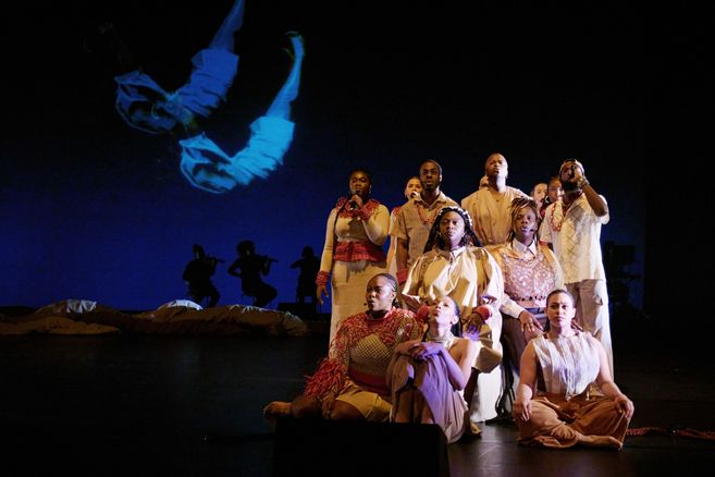 A group of performers in beige and red costumes sit and stand in formation on stage. In the background, silhouettes of musicians and a projection of a floating body are visible.