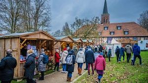 Menschen besuchen einen Weihnachtsmarkt mit Holzhütten und einer Kirche im Hintergrund.