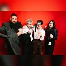 Vier Personen posieren fröhlich vor einem roten Hintergrund. Eine Person hält eine Flasche, eine andere trägt eine Krawatte um den Kopf.