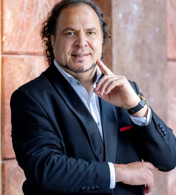 Ein Mann in einem schwarzen Anzug lehnt an einer Wand und lächelt in die Kamera.