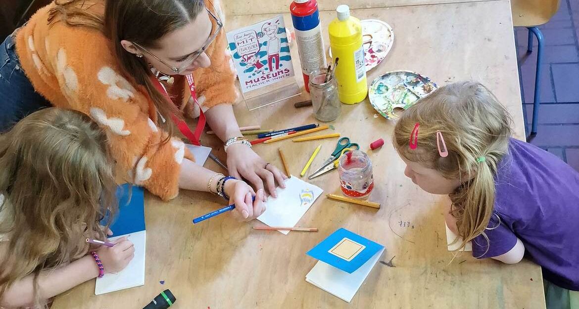 Erwachsene und zwei Kinder basteln an einem Tisch mit Farben und Papier.