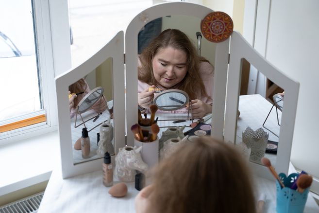 Eine hellhäutige, junge Frau sitzt an einem weißen Schminktisch, auf dem ein großer, dreiteiliger Spiegel montiert ist. Der Tisch steht vor einem Fenster.  Auf dem Schminktisch stehen verschiedene kosmetische Produkte, die für ein Make-up benötigt werden – z.B. Schwämmchen, Pinsel und Puder. Die verschiedenen Pinsel stecken in dekorativen kleinen Bechern. Die Frau hält einen Mascara-Pinsel und einen Kajalstift in der Hand. Sie ist offenbar dabei, ihre Augen zu tuschen.  Das Foto ist vom Rücken der Frau aus aufgenommen. Man erkennt ihr konzentriertes Gesicht von vorne im großen mittleren Spiegel des Schminktischs. Sichtbar ist in dem großen Spiegel auch, dass vor ihr auf dem Tisch zwischen den Schminksachen auch noch ein kleiner runder Aufstellspiegel steht, auf den sie gerade schaut. Hinter ihrem Rücken erkennt man den Griff ihres Rollstuhls.  Die Frau ist etwa 25 Jahre alt, sie hat dunkelblonde Haare, die offen auf ihren Schultern liegen. Sie hat ein rosa Kapuzen-Sweatshirt an.