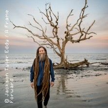 Person mit langen Dreadlocks steht am Strand vor einem kahlen Baum.