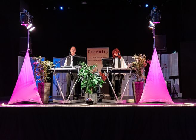 Zwei Musiker auf einer Bühne mit Keyboards und Mikrofonen, umgeben von Pflanzen und pinkem Licht.