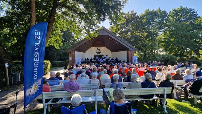 Publikum sitzt bei einem Benefizkonzert in der Musikmuschel Bad Bodenteich; auf der Bühne spielt eine Blaskapelle, links steht ein blaues Banner der Bundespolizei.