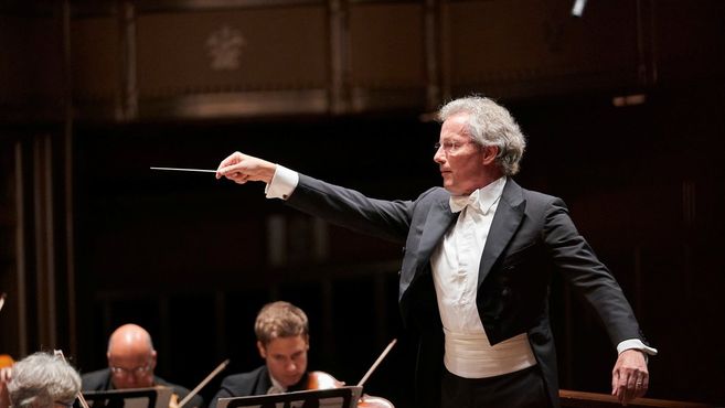 Ein Dirigent in Frack dirigiert ein Orchester auf einer Bühne.