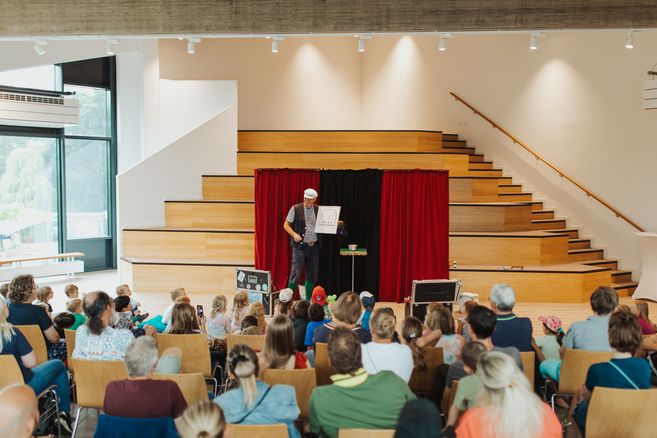 Ein Zauberer in Matrosenkleidung führt vor einem Publikum von Kindern und Erwachsenen auf einer Bühne mit roten Vorhängen eine Show auf.